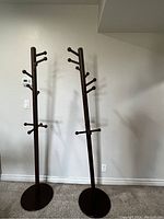 Two brown wooden freestanding coat racks with 11 hooks each and round solid bases against a light wall.