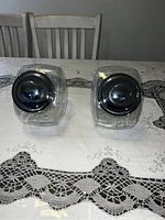 Two clear glass jars with silver lids placed on a white lace tablecloth in front of two wooden chairs, shown from the bottom side to display lids and jar shape.