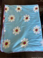 Mini wool blanket laid flat, showing light blue color with white daisies and red/white stitched edges