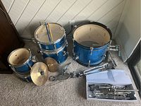 Blue drum shells and cymbals laid on carpet with metal stands and parts boxed next to them.