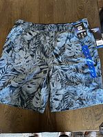 Front view showing the swim shorts laid flat, displaying the elastic waistband, side pockets, blue leaf pattern, and size tags.