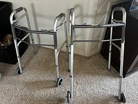 Photo shows two metal walking frames with silver finish, front wheels, adjustable height, and hand grips, standing side by side on carpeted floor.