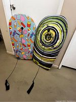 Two water boards leaning against a wall, one with food-themed colorful patterns and the other with a yellow-black swirl design, both with attached safety handles