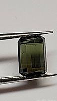 Photo of a rectangular octagon cut green tourmaline gemstone held by tweezers against a white background, showing front facets and color.