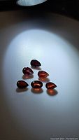Seven cherry red garnet gemstones in pear and oval cuts on white background under light showing facets and color.