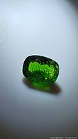 Photo showing cushion cut natural chrome diopside gemstone with deep electric green color, placed on a light surface for visibility of color and inclusions.
