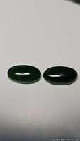 Two deep green oval jade cabochons displayed on a white background, showing smooth polished surfaces and elongated oval shapes.