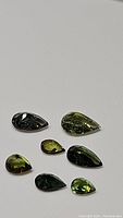 Seven pear-shaped green tourmaline gemstones laid out on a white surface, showing variations in green color and internal inclusions.