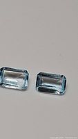 Two natural emerald cut sky blue topaz gemstones placed side-by-side on a plain light background, showing their size and color.