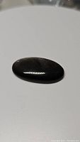 Polished oval cabochon silver sheen obsidian gemstone lying flat on white surface showing dark base color without visible silver sheen flash.