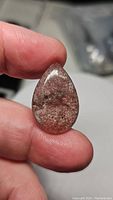 Close-up photo of a pear-shaped natural strawberry quartz cabochon held between fingers showing reddish-pink internal inclusions and polished surface.