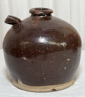 Front view of antique brown glazed ceramic jug with small spout and unglazed rim base.