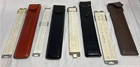 Five slide rules with leather pouches arranged in a row showing size variation and storage cases.
