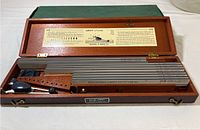 Open wooden box showing metal letter templates arranged inside with two black drafting pointers and a wooden protractor tool. Instruction sheet visible inside the lid.