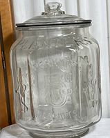 Front and side view of clear vintage Planters Peanuts jar with embossed text and Mr. Peanut figures, showing lid and metal rim with some aging and small chips.