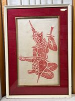 Full view of framed red stone rubbing print depicting a seated figure with intricate traditional Siamese attire and sword, with red mat and white frame.
