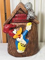 Front and slight side view of the ceramic Woody Woodpecker cookie jar showing the character climbing out of a treehouse with a squirrel on the roof.