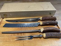 Four-piece carving set laid out on a wooden surface with cream case closed behind.