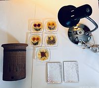 Mixed vintage lot arranged on white surface showing pottery vase, gooseneck lamp, two enamel trays, and five dried flower coasters with holder.