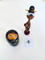 Photo of two Japanese Kokeshi dolls side by side on white background. The mother and child doll is rounded with floral painted details, the contemporary doll is slender with smaller child on head.