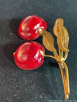 Front view of the brooch showing two red glass cherries and gold-tone metal leaves and stems on a black background.