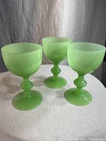 Three vintage jadeite Fenton juice cups, displayed upright on a textured white surface showing the front view of the cups and their green opaque glass with bulbous stems.