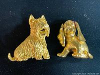 Two vintage gold tone dog brooches, a Westie marked Trifari and a spaniel marked Monet, displayed on black background.