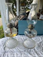 Both antique kerosene lamps displayed side by side on a white cloth, showing glass bases and chimneys