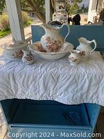 Photo showing the entire antique wash basin set displayed on a white cloth, including large pitcher, large basin, small pitcher, two small bowls, and covered sugar bowl, all with brown floral decoration on white ceramic.
