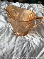 Pink carnival glass creamer with a floral and linear embossed pattern and an iridescent finish on fabric background