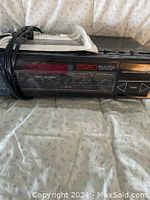 Front angled view of the black stereo clock radio showing buttons, display, and power cord coiled on top, resting on patterned fabric.