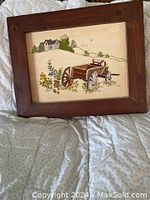 Photo showing the framed needlework with a country scene including a wooden cart, flowers, a house on a hill, and trees.