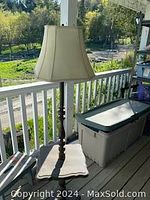 Floor lamp with wooden turned base, square table integrated on the base, and beige fabric shade captured outdoors on a porch with white railing and greenery in background.
