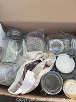 Box of assorted glass storage jars visible with clip-on lids and white gasket seals. Jars are of different sizes and shapes, mostly cylindrical. Some are packed with bubble wrap and fabric storage bags.