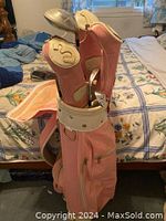 Golf bag standing upright on bed showing pink color, several zippered compartments, and golf club shafts inside.