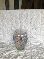 Oval-shaped clear glass vase with colorful hand painted flowers placed on light patterned fabric background.