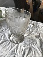 Front angle view of the large clear glass pitcher showing embossed floral daffodil and vertical stripes with footed base and handle