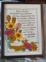 Framed needlework in brown wooden frame, colorful embroidery showing flowers, utensils, pie and poem text, 11x13 inches