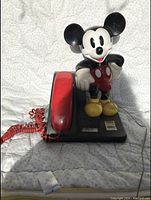 Front view of vintage Mickey Mouse novelty telephone. Shows standing Mickey Mouse figure with red handset on black base and coiled red phone cord.