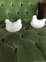 Two white opaque milk glass hen on nest figurines displayed on a green tufted chair.