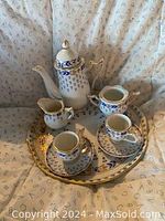 Miniature porcelain tea set displayed on patterned fabric background. Set includes teapot with lid, sugar bowl with lid, creamer, two teacups and saucers, on a round tray. Blue floral hand-painted pattern with gold trim visible on all pieces.