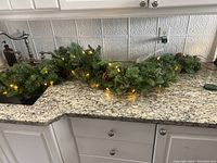 Full view of the green artificial garland with integrated warm white lights and pinecones laid out on a kitchen countertop.