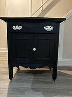 Front view of black antique washstand with drawer and double doors, white handles and knob, on casters.