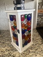 Front and side view of the white lantern showing colorful circular stained glass stones attached to the frame. Lantern has a small black metal handle on top.