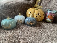 Five items displayed: an orange pumpkin with cutout designs; three small ceramic pumpkins in colors gray, white, and blue-green with gold words 'grateful', 'thankful', 'blessed'; and a red jar candle labeled Ashland Apple Orchard.