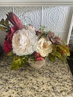 Front view of custom faux floral arrangement showing white peony-like flowers, dark red roses, greenery in a white ceramic vase on granite countertop.