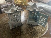 Pair of iron candle holders on decorative table, showing their overall shape, embossed top and cutout panels.