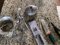 Wide view of kitchen tools displayed on granite countertop including Moulin Legumes, hand beater, veggie steamer, knives, and battery operated meat fork.