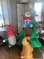 Photo showing the inflatable Santa climbing a decorated Christmas tree with an orange inflatable dog at the base, next to a separate inflatable elf figure.