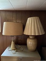 Photo showing the pair of lamps on a surface, one tall ceramic with pleated shade, one shorter wooden with drum shade.